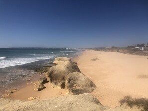 A beach with clear skies, rocky formations, and a distant shoreline with buildings. - Refúgio Ls (Guia)