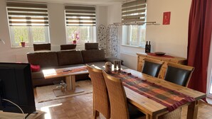 Una sala moderna con un sofá, una mesa de comedor y sillas. Hay ventanas con persianas, un radiador y un televisor.