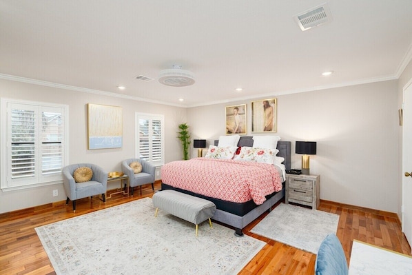 A bedroom with a large bed, two armchairs, a bench, a nightstand, and a window with blinds.