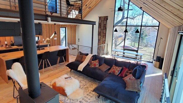 A spacious loft-style living area with a large dark-colored sofa, a fireplace, and a view of the outdoors through large windows. - This deluxe modern new 3-story home with central AC offers the best of Bozeman! (Bozeman)