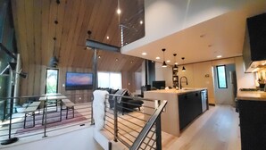 A spacious living area with a high ceiling, wooden beams, a dining area with a table and chairs, a kitchen with a central island, and a TV mounted on the wall. - This deluxe modern new 3-story home with central AC offers the best of Bozeman! (Bozeman)