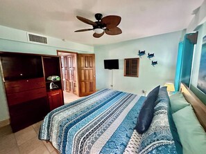 Un dormitorio con cama, armario, ventilador de techo, televisión y puerta que da a otra habitación.