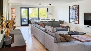 Un salon moderne avec un canapé gris, une table basse et un téléviseur. Il y a une grande fenêtre donnant sur des arbres et un balcon.