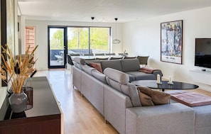 A modern living room with a grey sofa, a coffee table, and a TV. There's a large window with a view of trees and a balcony.