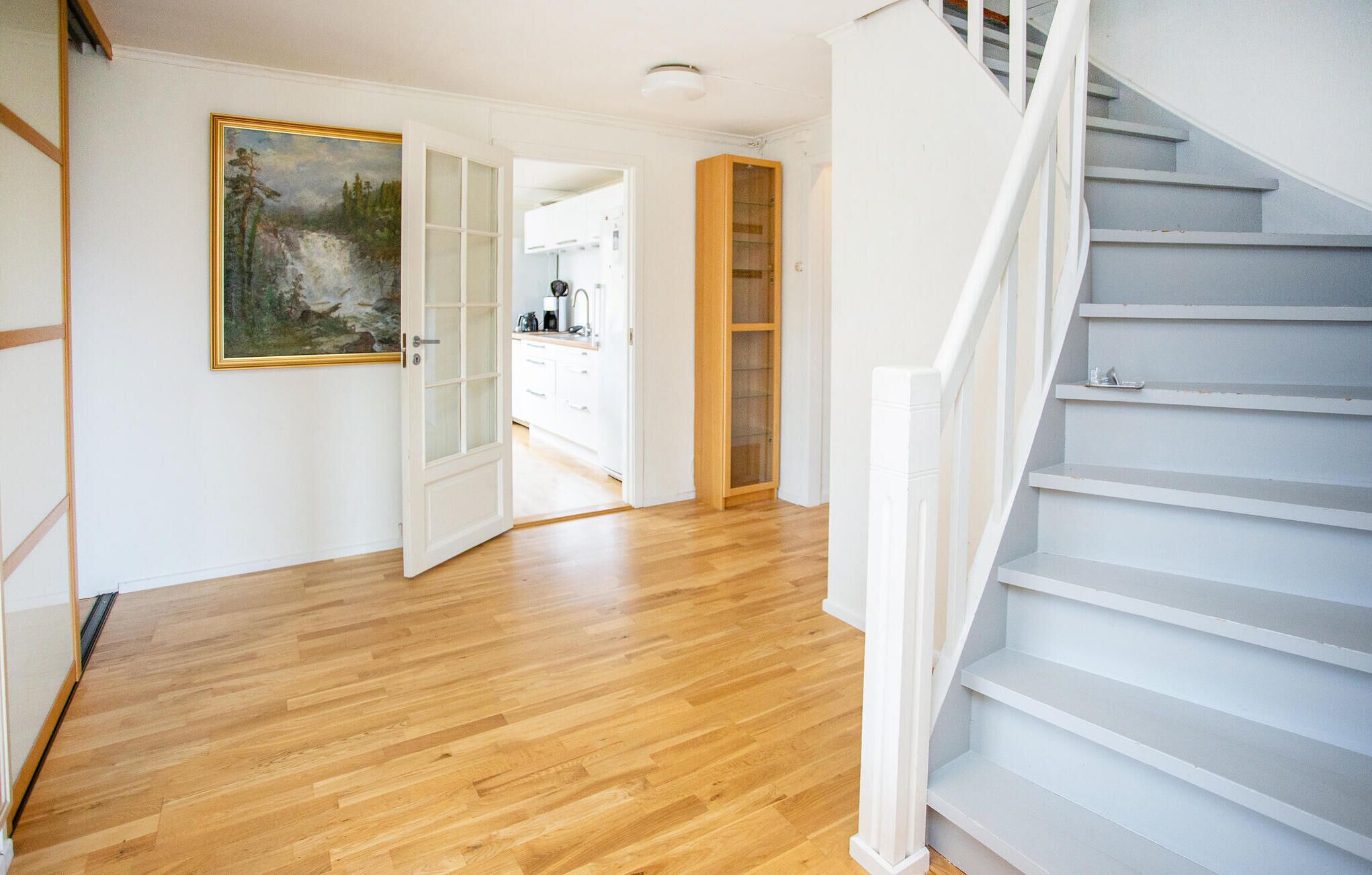 A spacious hallway with wooden flooring, a white staircase, and a kitchen area visible through an open door.