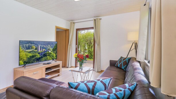 A modern living room with a leather sofa, a flat-screen TV, a small table with a vase of flowers, and a view of a garden through an open door. - Ferienhaus 4 in Kirchheim (Kirchheim)