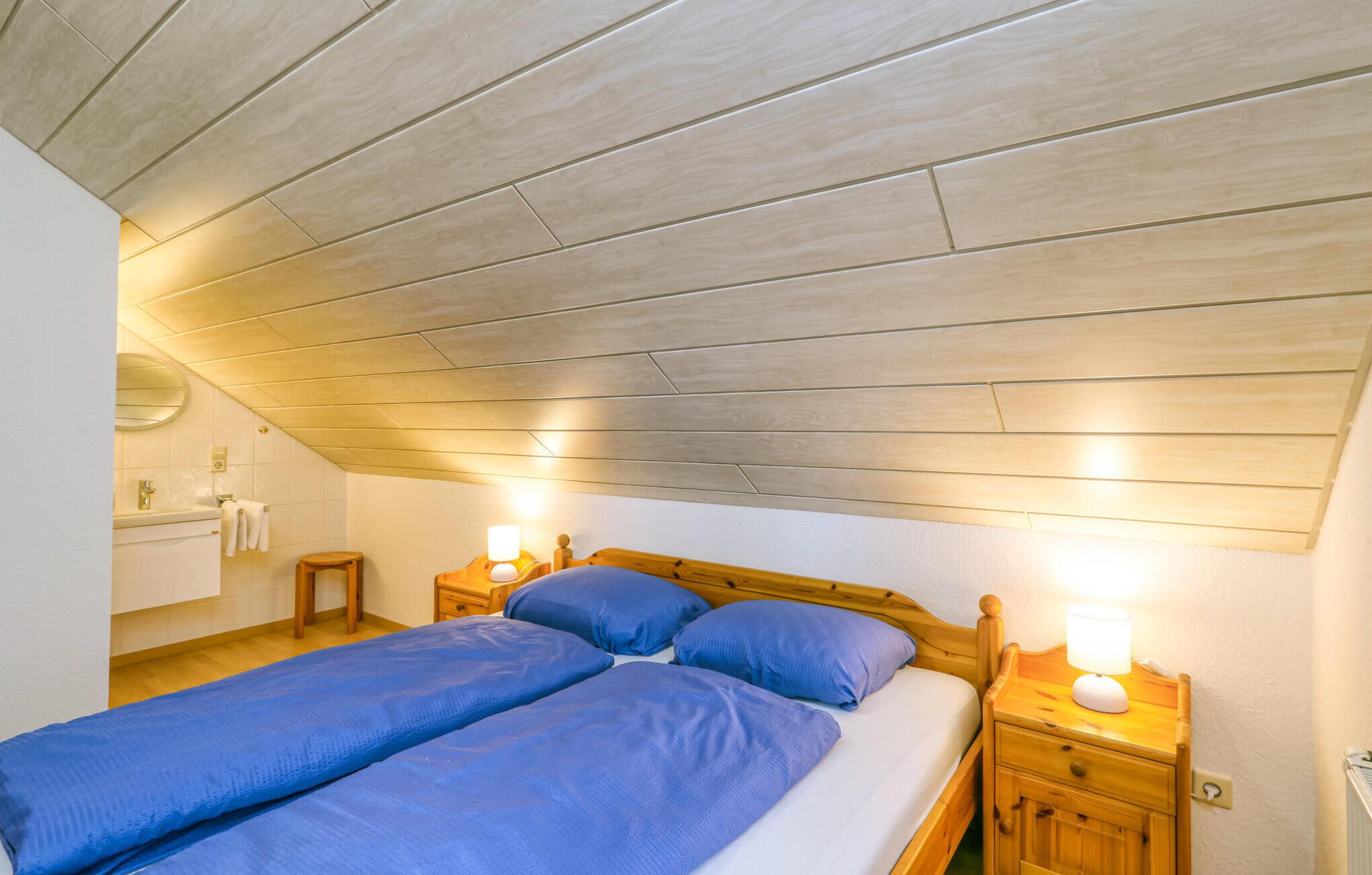 Ein Schlafzimmer mit schräg versiegelter Decke, einem Doppelbett mit blauer Bettwäsche, zwei Nachttischen mit Lampen, einem kleinen Holzschemel und einem runden Spiegel über einem weißen Waschbecken.