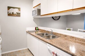 Una cocina con gabinetes blancos, encimera de granito, pileta de acero inoxidable, microondas y un reloj en la pared.