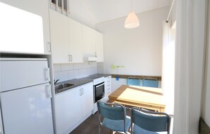 Ett kompakt kök med vita skåp, en inbyggd ugn, en diskho och en matplats med ett träbord och blå stolar.