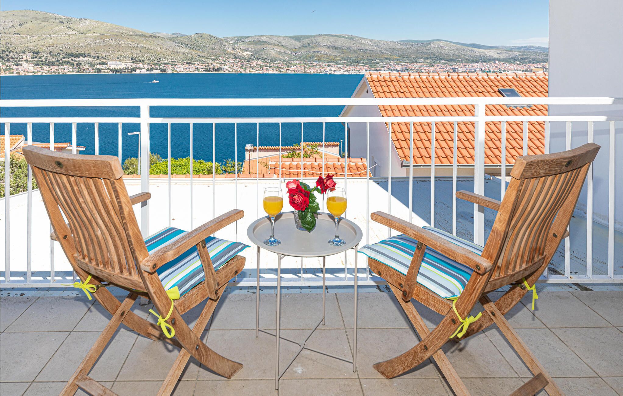 A balcony with two wooden chairs and a small table, overlooking a coastal town and the sea.