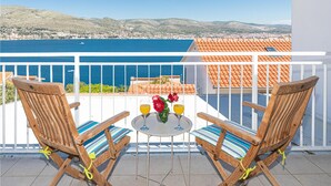 A balcony with two wooden chairs and a small table, overlooking a coastal town and the sea.
