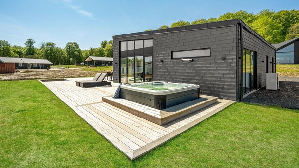 A modern outdoor hot tub area with a wooden deck, surrounded by a well-maintained lawn and a brick building.