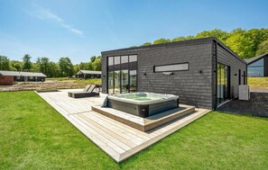A modern outdoor hot tub area with a wooden deck, surrounded by a well-maintained lawn and a brick building.