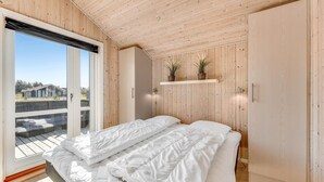 Une chambre avec un grand lit, des murs en bois, une porte-fenêtre coulissante donnant sur un balcon et deux plantes en pot posées sur une étagère.