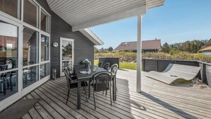 Une terrasse extérieure couverte, équipée d’une table et de chaises, un petit jardin et une vue sur une maison dont le toit est recouvert de tuiles rouges.