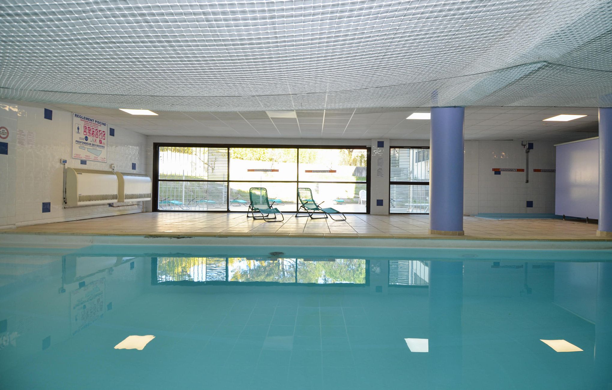Piscina interna com paredes de vidro transparente, piso ladrilhado e várias cadeiras de descanso.