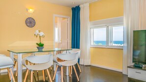 Un comedor moderno con una mesa de vidrio, sillas blancas, una orquídea en maceta y vista al mar a través de amplias ventanas.