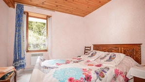 Een slaapkamer met een houten plafond, een bed met bloemenbeddengoed, een raam met uitzicht op bomen en een stoel.