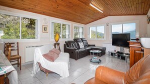 Een ruime woonkamer met een bank, een eethoek met stoelen, een televisie en een groot raam met uitzicht op het omliggende landschap.