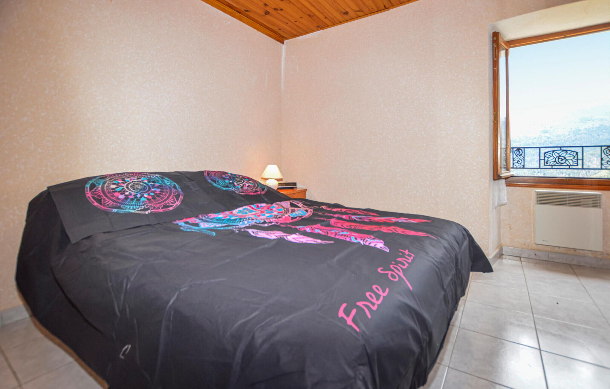 A bedroom with a bed covered in a black bedspread with colorful patterns, a nightstand with a lamp, and a window with a view of a snowy landscape.