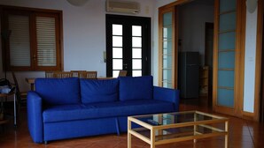 A living room with a blue sofa, a glass coffee table, and a kitchen area visible through an open door. - Holiday flower villa with pool (Misterbianco)