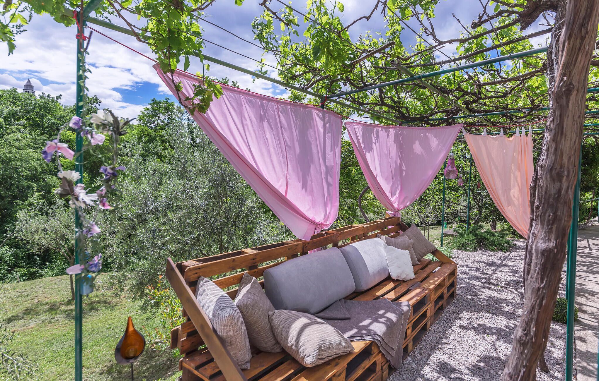 A wooden bench with cushions and a hanging pink cloth structure in a garden setting.