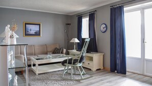 A living room with a sofa, a glass coffee table, a rocking chair, a side table with a lamp, a framed picture on the wall, and a large window with curtains.