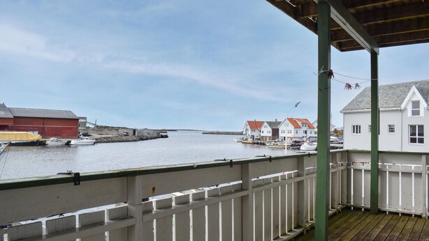 A waterfront view from a covered balcony with a wooden deck, overlooking a calm harbor with several boats and buildings.