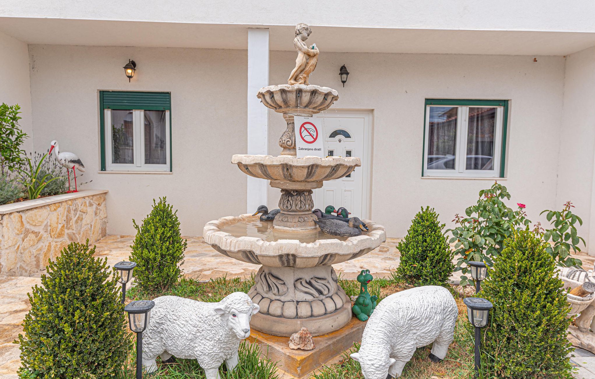 Ein Brunnen mit Enten, umgeben von kleinen Bäumen und einer Schafstatue.
