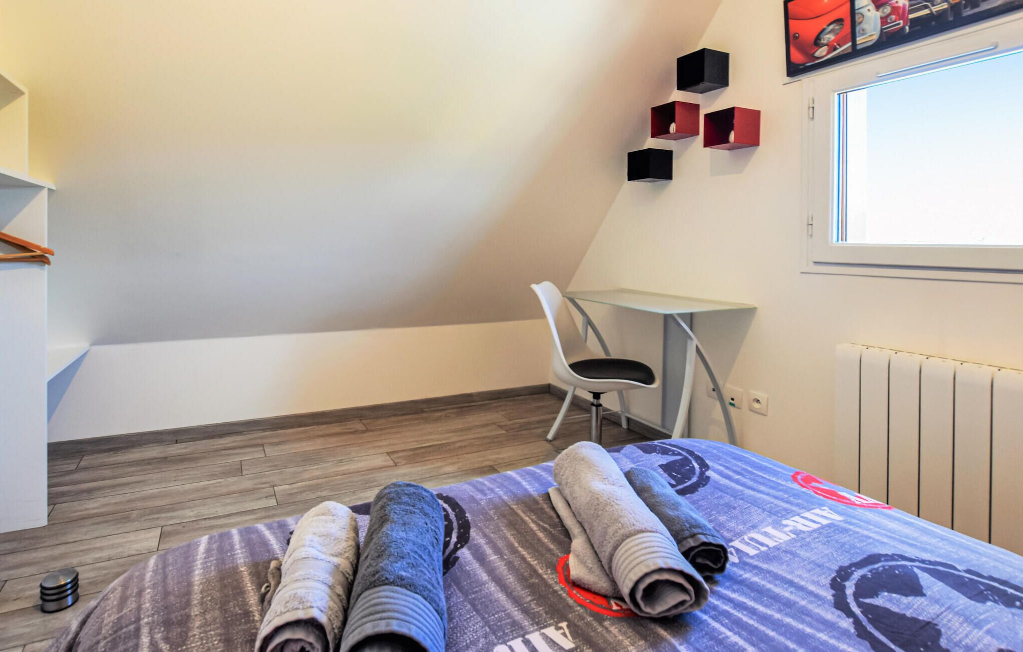 Una habitación pequeña y moderna con una cama, una silla y una mesita. Hay toallas sobre la cama y una ventana con vistas al exterior.