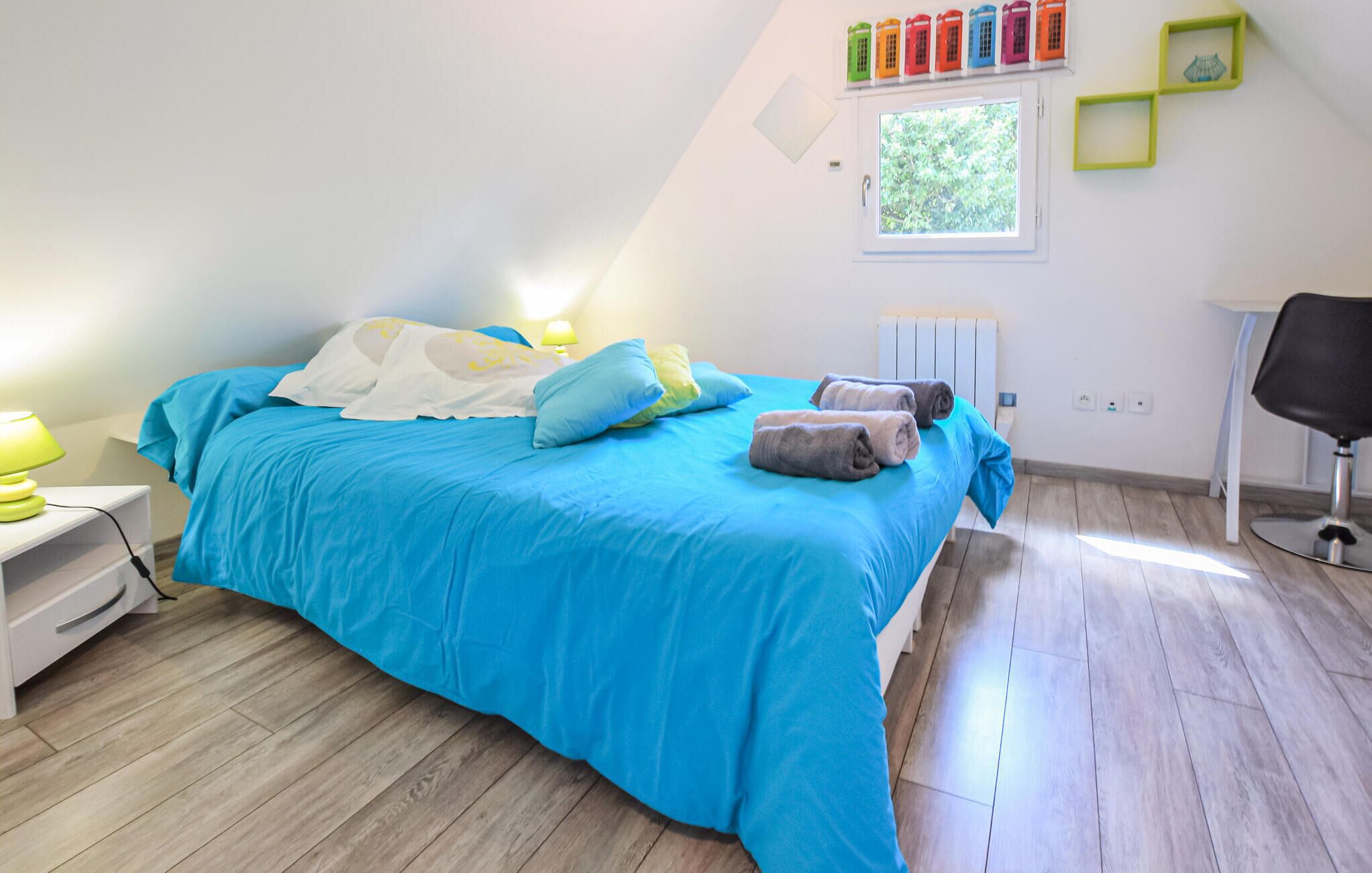 A bedroom with a blue bedspread, a white nightstand with a lamp, a radiator, and a window with a view of trees.