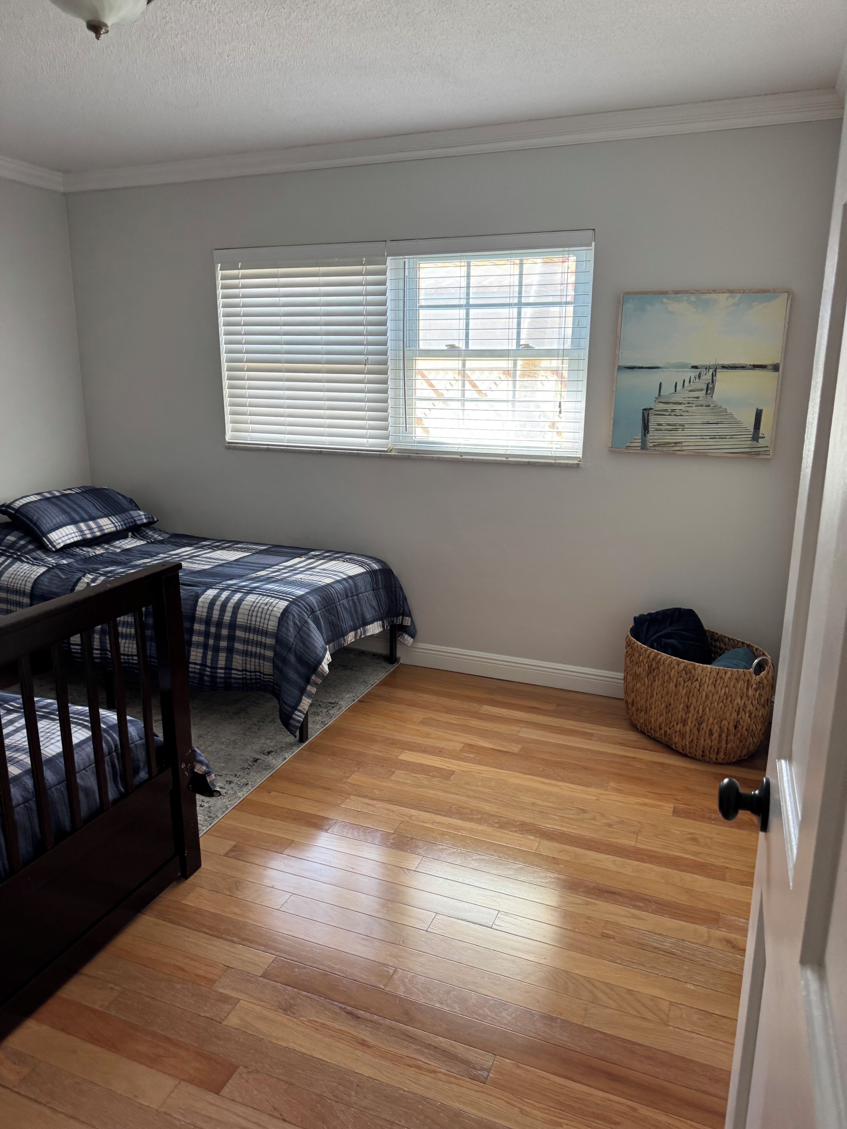 Une chambre avec deux lits, un plancher en bois, une fenêtre avec des stores, un tableau au mur et un panier près de la porte.