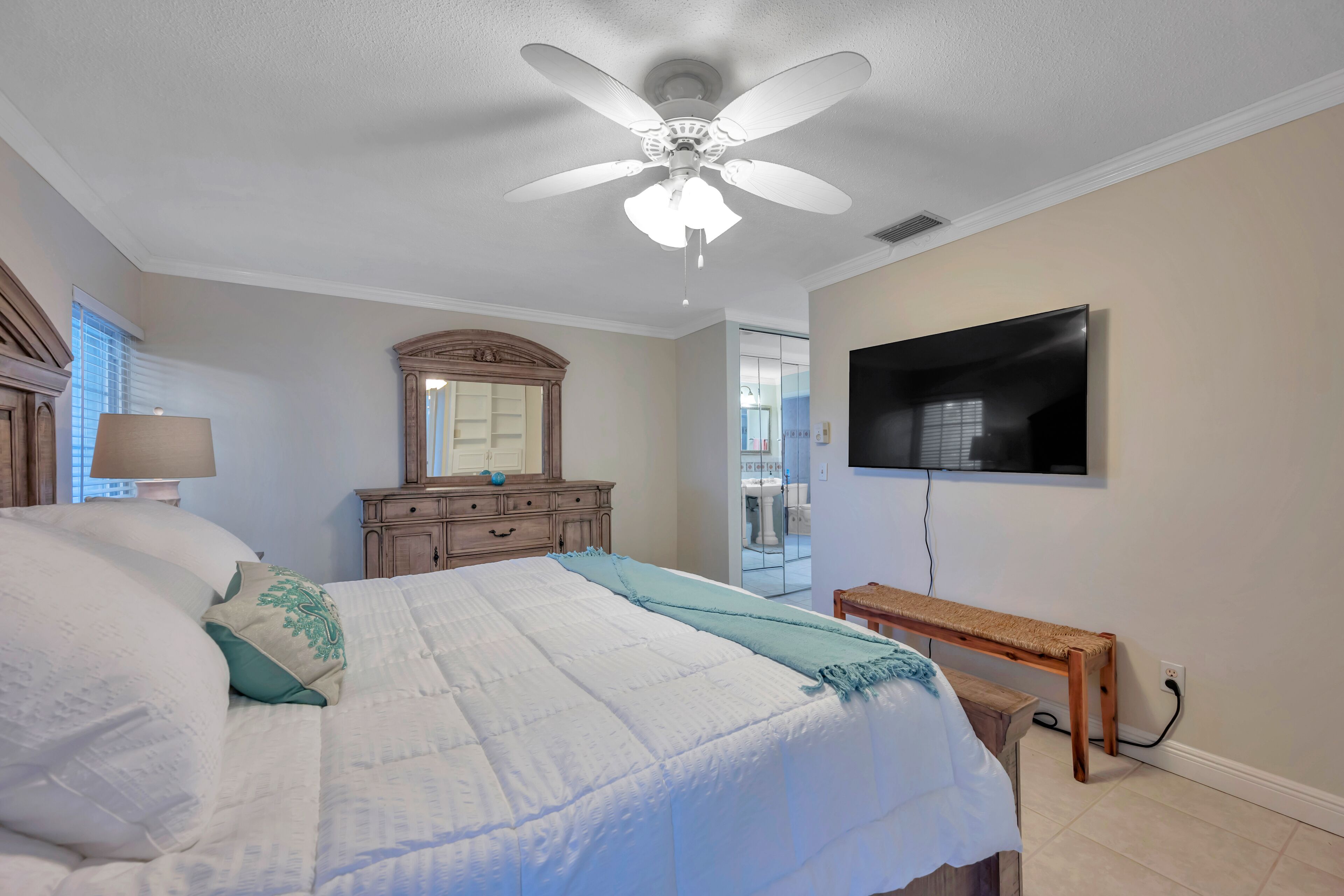 A bedroom with a bed, a ceiling fan, a dresser, a mirror, a television, and a bench.