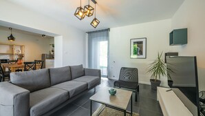 Un saló modern amb un sofà gris, una taula de centre i una televisió. Al fons, hi ha una zona menjador amb una taula de fusta i cadires.