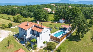 Una casa moderna de dos pisos amb piscina, envoltada de vegetació exuberant i un paisatge de muntanyes.