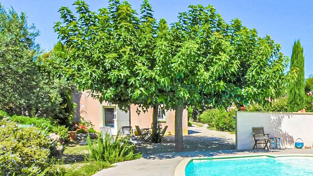 Ett poolomr åde med en klarblå pool, omgivet av frodig grönska och en byggnad med en beige fasad.
