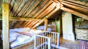 Een knus zolderkamer met houten plafond, een bed met witte beddengoed, een kleine houten trap en een dressoir met lades.