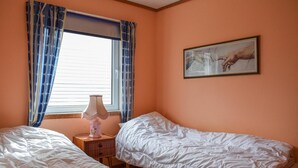 Et soveværelse med to senge, et vindue med blå gardiner, et natbord med en lampe og et billede på væggen.