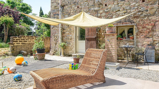 Une chaise en osier placée sous un auvent, un bâtiment en pierre avec une porte, et des plantes en pot.