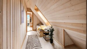 Un intérieur en bois avec un plafond en pente, un lit recouvert d’une couverture jaune, une plante en pot et un tapis à motifs géométriques.