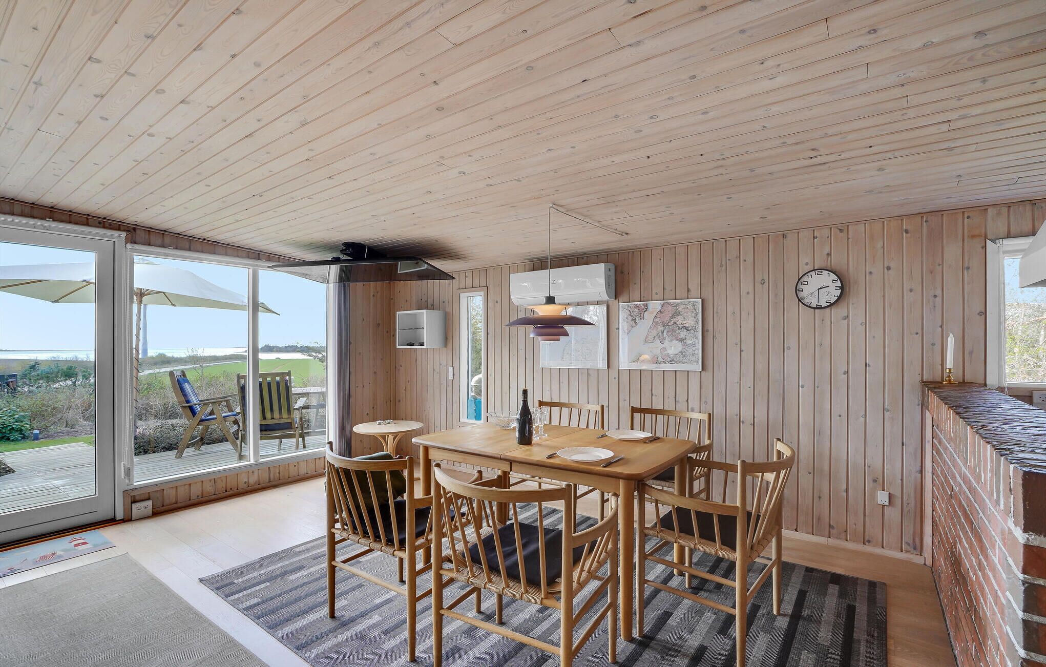 Een eethoek met een houten tafel en stoelen, een hangklok en een groot raam met uitzicht op de omgeving.