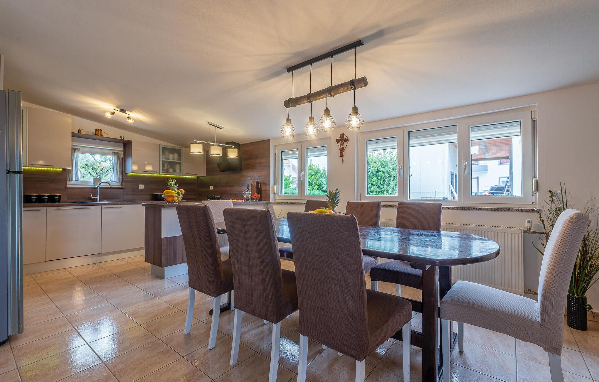 A modern kitchen with a dining area, featuring a central island, pendant lighting, and large windows.