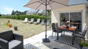 Un patio al aire libre con una mesa, sillas y un paraguas. Se ven plantas en macetas y un seto al fondo.