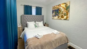 Un lit bien fait, avec une tête de lit grise, des oreillers blancs et une couverture beige. On y trouve une petite table de chevet avec une lampe, et un tableau représentant une scène de rue, accroché au mur.