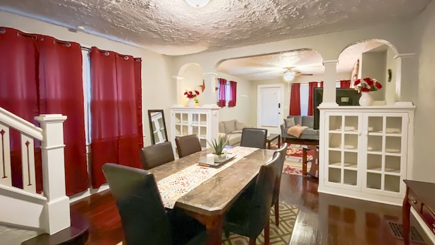 Un coin repas avec une table en bois, des chaises noires et un tapis à motifs. On aperçoit en arrière-plan un meuble blanc aux portes vitrées et un rideau rouge.