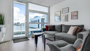 Ein modernes Wohnzimmer mit einem grauen Sofa, einem kleinen runden Tisch und Blick auf einen Hafen durch große Fenster.