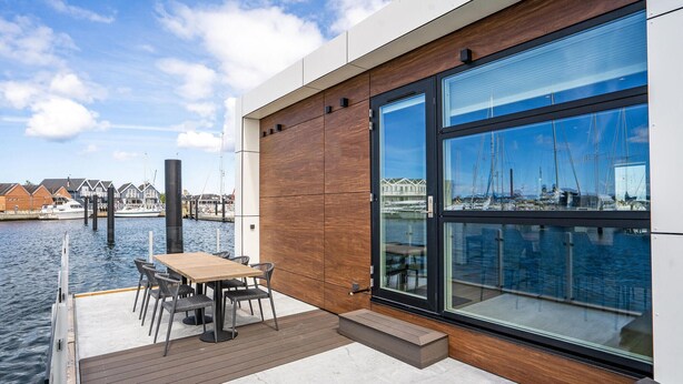 Eine moderne Terrasse im Freien mit Holztisch und -stühlen, mit Blick auf einen Yachthafen, wo Boote und ein Holzgebäude im Hintergrund zu sehen sind.