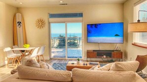 A living room with a large flat-screen TV, a sofa, a dining area with chairs, a surfboard, and a balcony with a view of the ocean. - ★★★★ Brand New Beautiful Ocean View Home ★★★★ (Whitethorn)