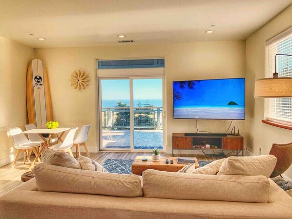 A living room with a large flat-screen TV, a sofa, a dining area with chairs, a surfboard, and a balcony with a view of the ocean.