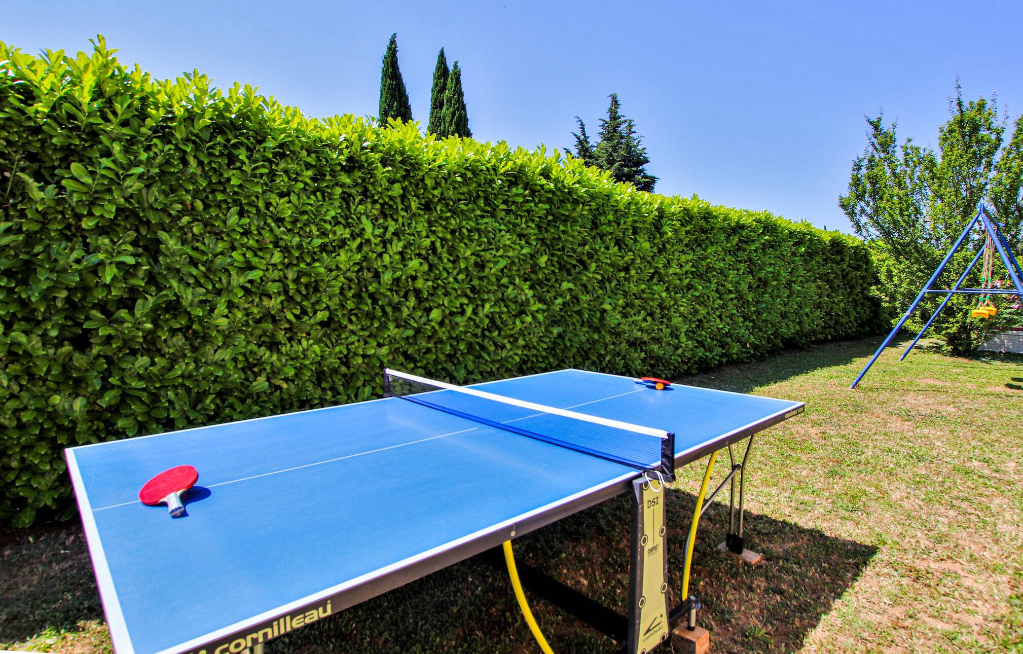 Mesa de ping pong com uma raquete vermelha, montada ao ar livre com uma sebe ao fundo.